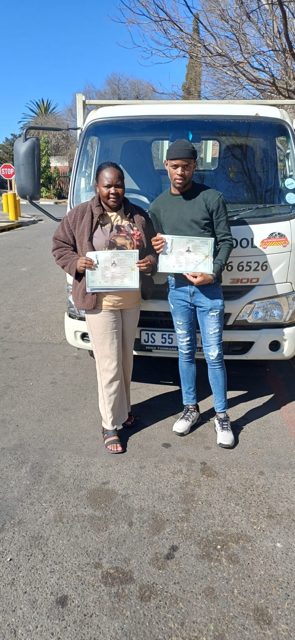 Two learners holding their licences in front of a 522 truck